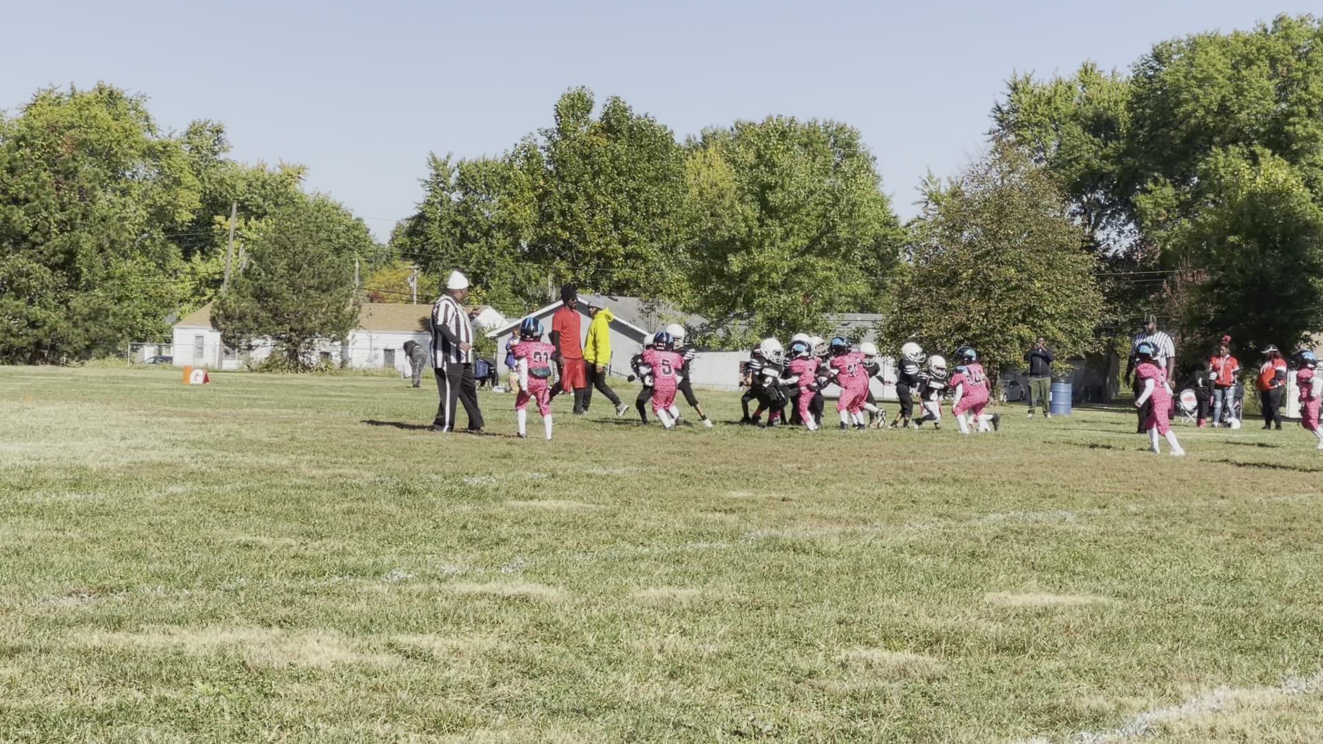 Youth football game. Players in pink uniforms kneel on a grassy field, referee stands nearby.