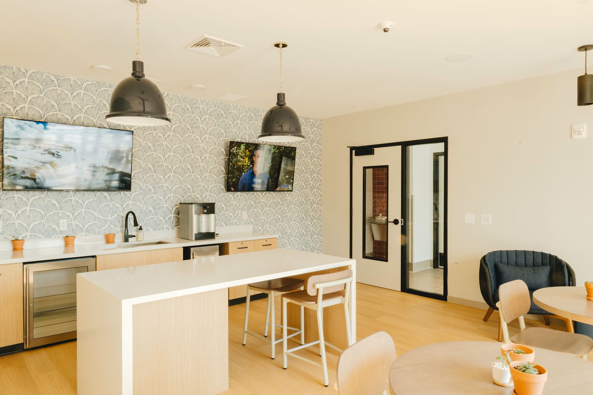 Modern lounge area with kitchen and seating; light wood floors, neutral walls, pendant lights, and two TVs at Revelry in Madison WI.