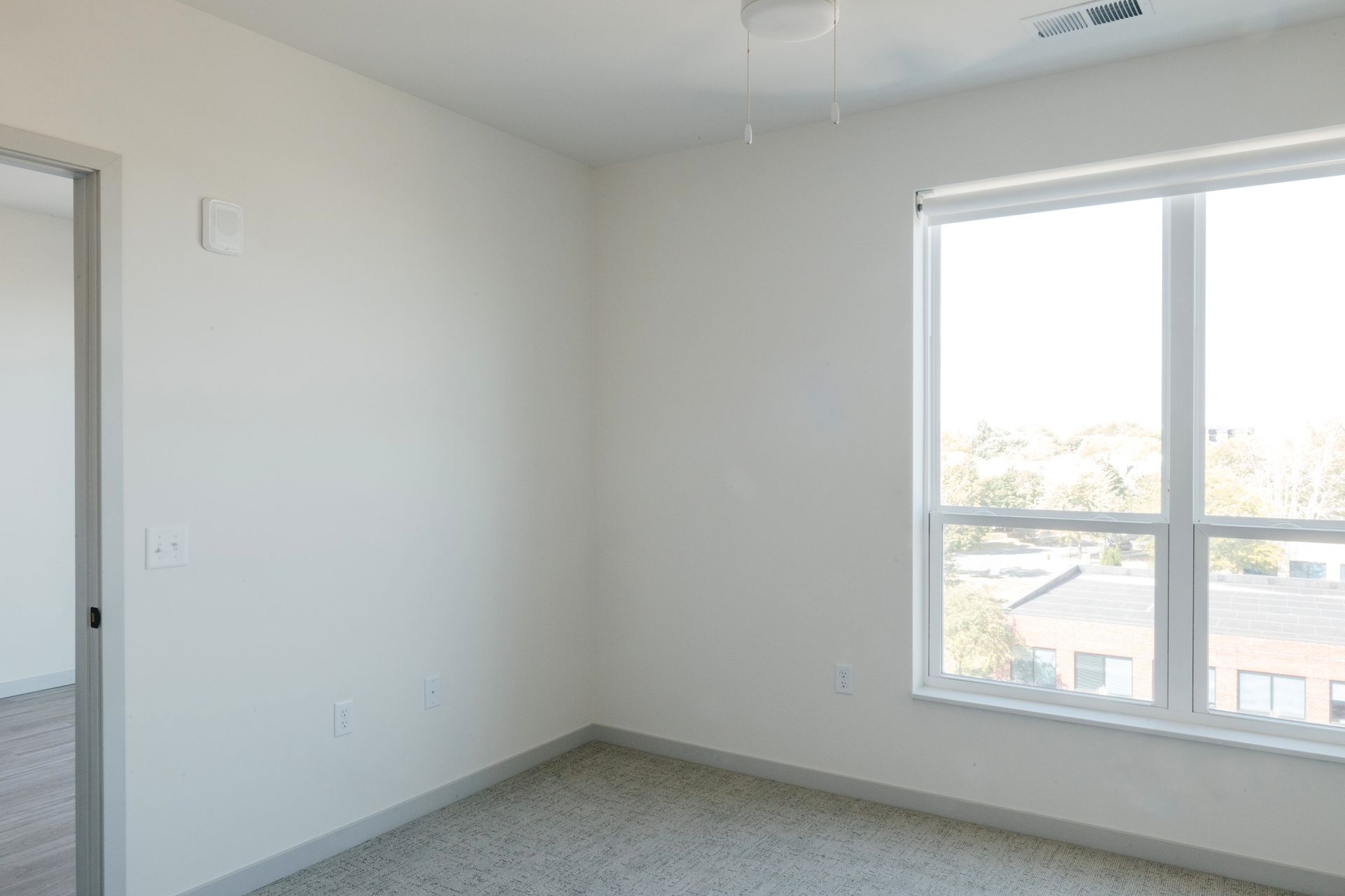 Empty, neutral-toned room with a window, door, and carpet. White walls and light gray carpet at Revelry in Madison WI.