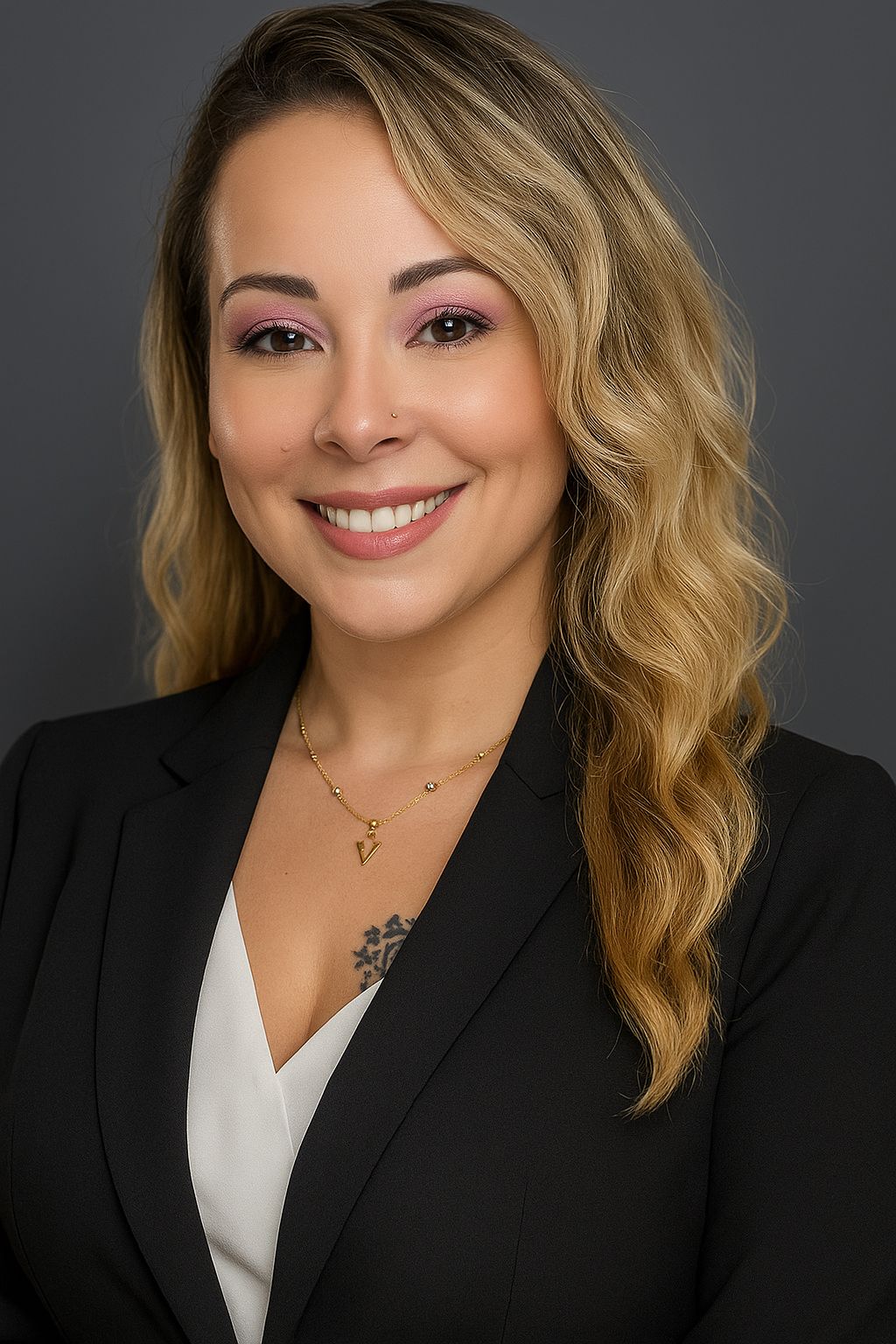 Woman in a black blazer, white top, and necklace, smiling. Gray background.