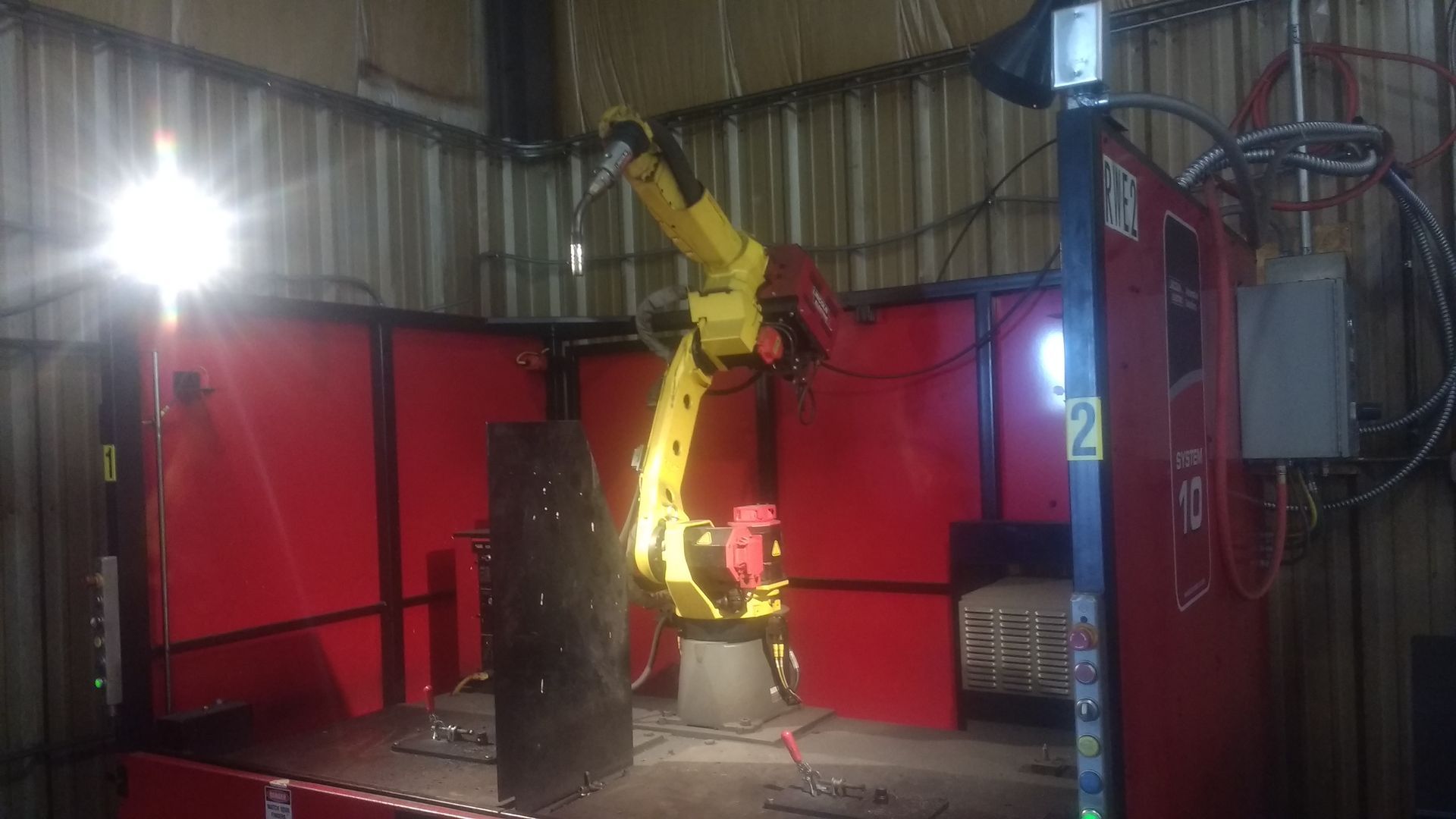 Yellow robotic arm welding in a red enclosed cell, bright light illuminates the scene.