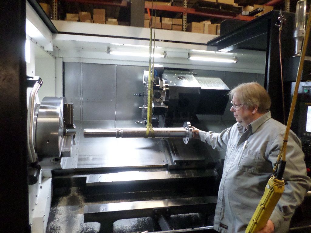 Man in work clothes holding a long metal rod near a large CNC machine in a workshop.