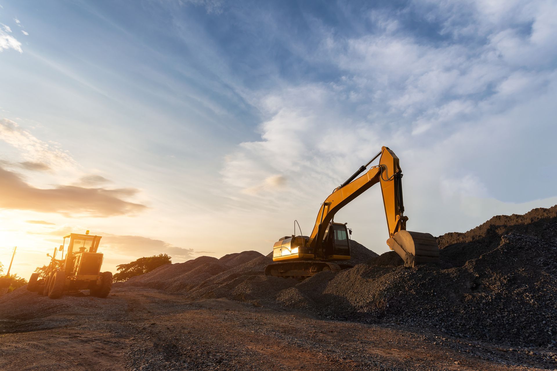 Construction site with excavator scooping rocks, bulldozer, and sunset sky.