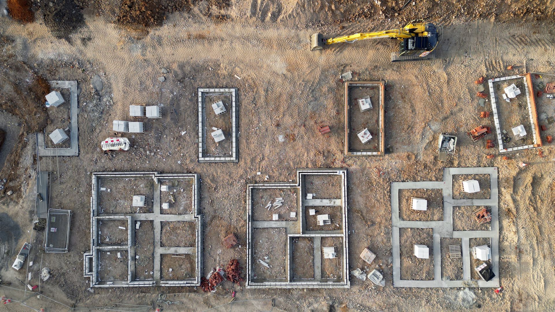 Overhead view of a construction site with concrete foundations, dirt, and a yellow excavator.