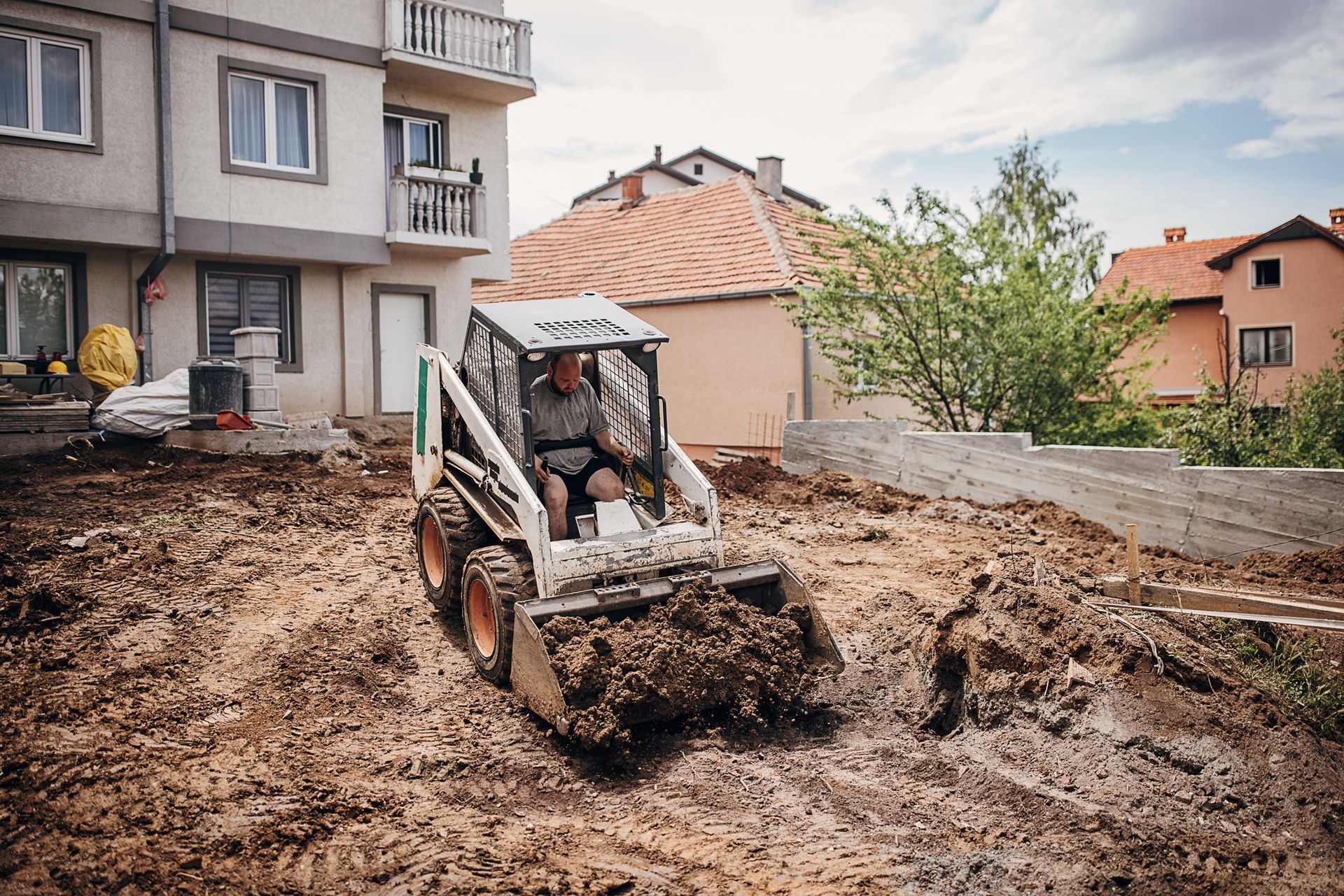 Man operating a skid-steer loader, moving dirt in a yard near a multi-story building.