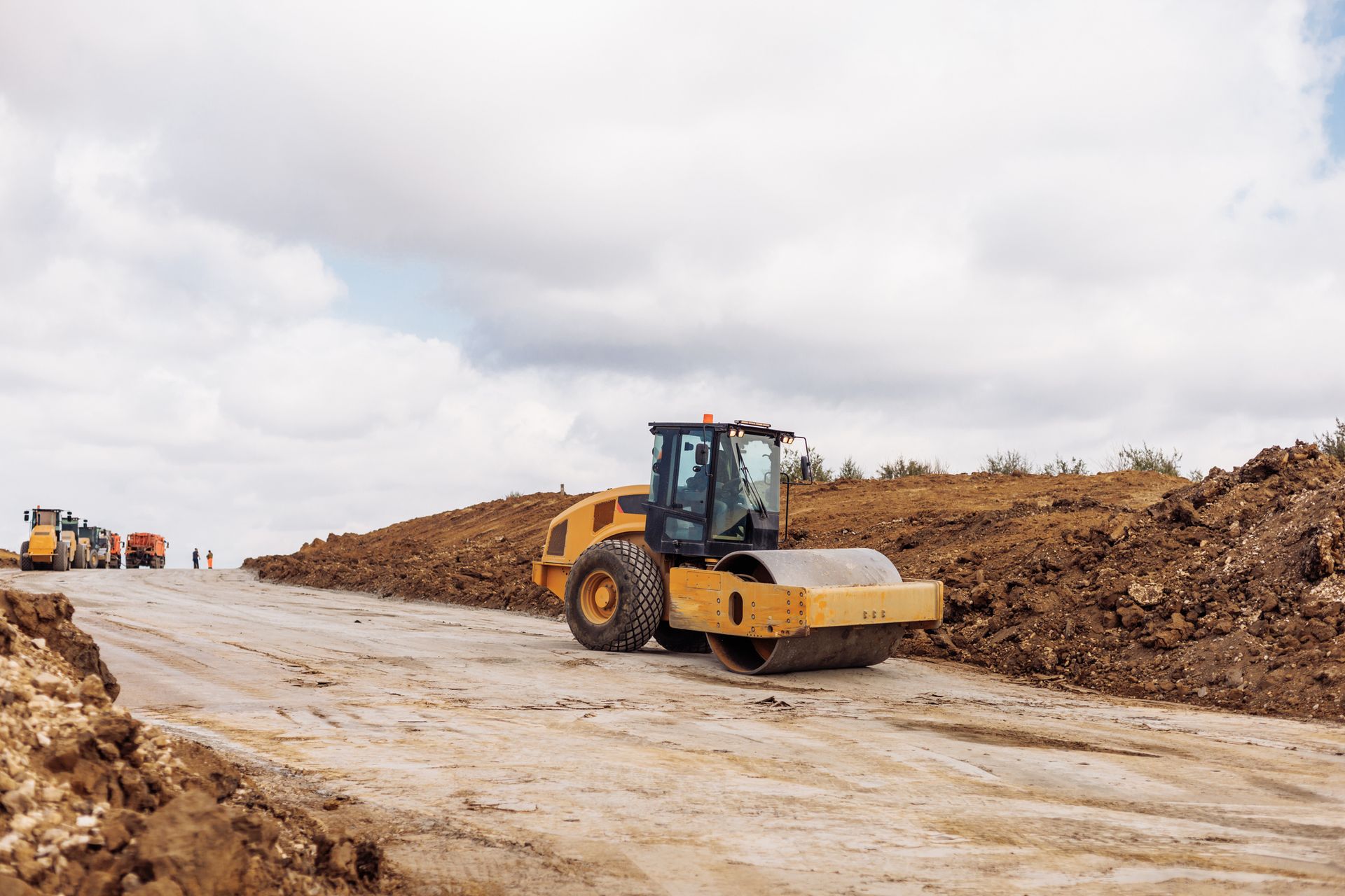 Yellow road roller compacting soil on a construction site under a cloudy sky.