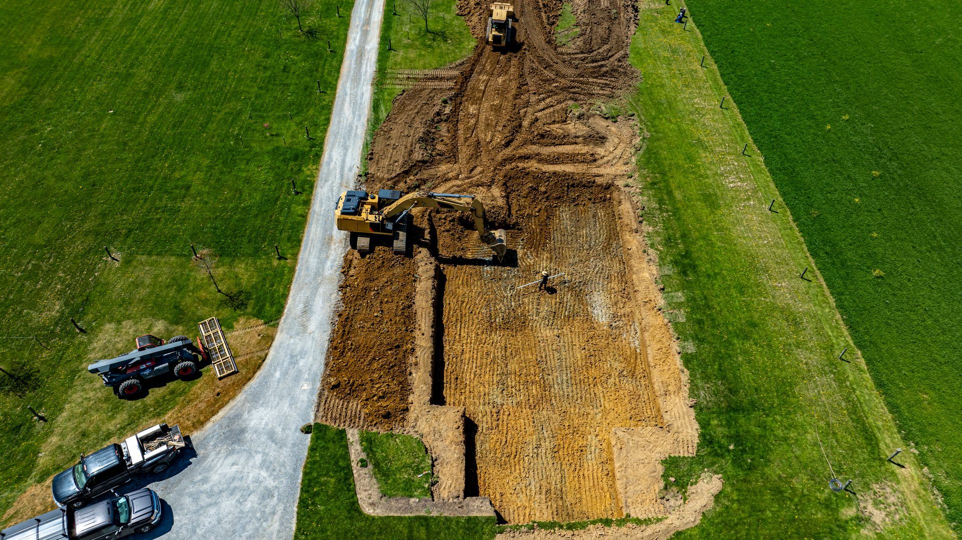 Construction site with heavy machinery on a dirt plot next to a grassy field and driveway.