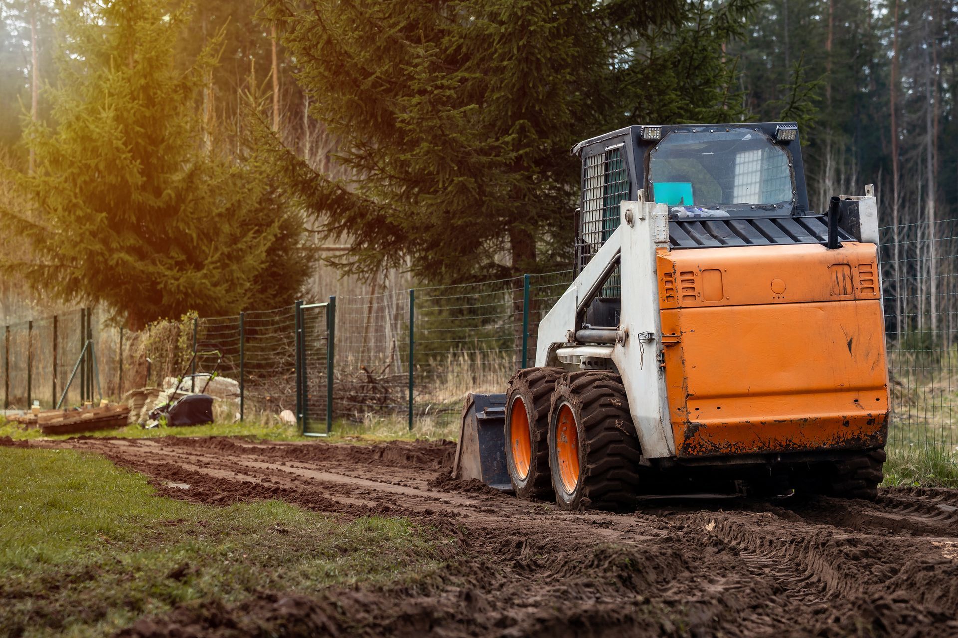 Orange and white skid steer operating on a muddy path near a forest with fence posts in the background.