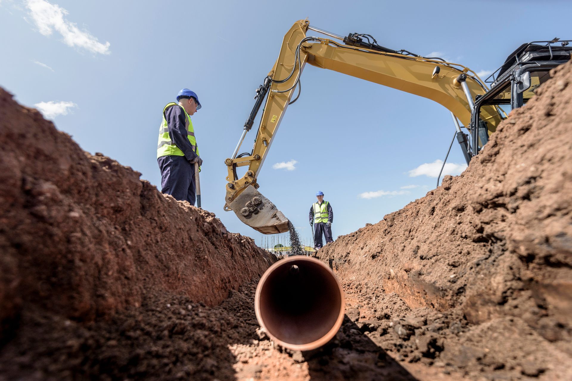 Workers installing a pipe in a trench with an excavator on a sunny day.