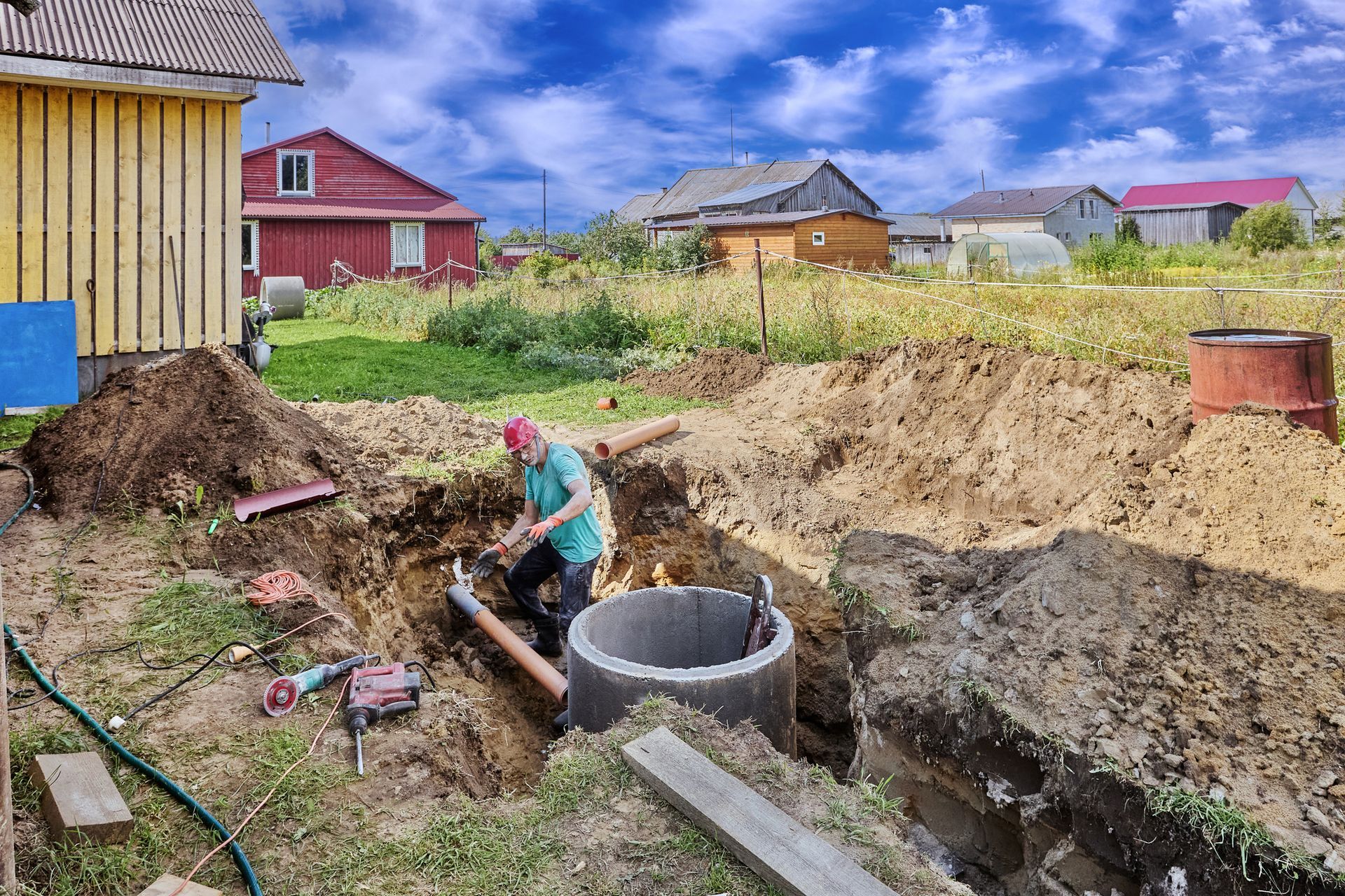 Man in a ditch working on plumbing near a concrete ring, with houses in the background on a sunny day.