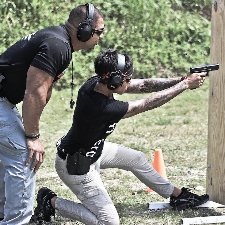 Man coaching a person kneeling, aiming a handgun at a target outdoors.