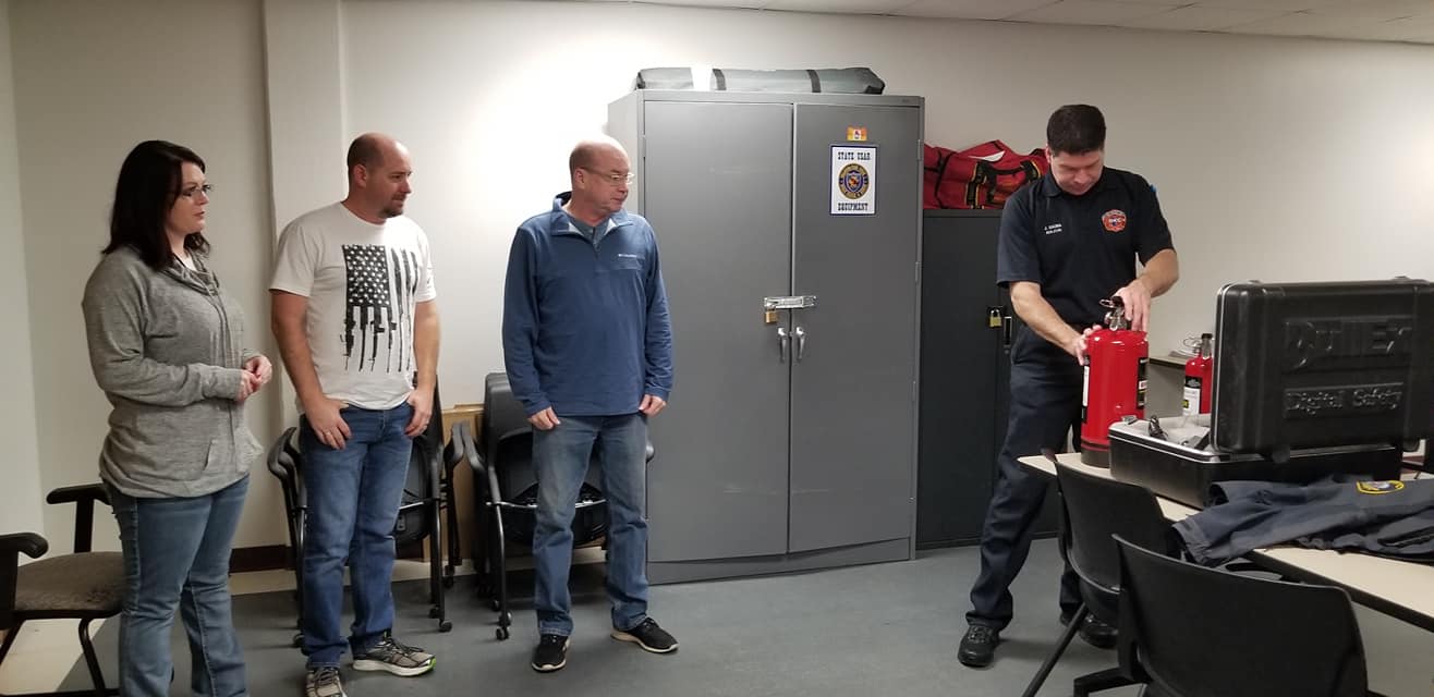 A person in uniform demonstrating a fire extinguisher for three people indoors.
