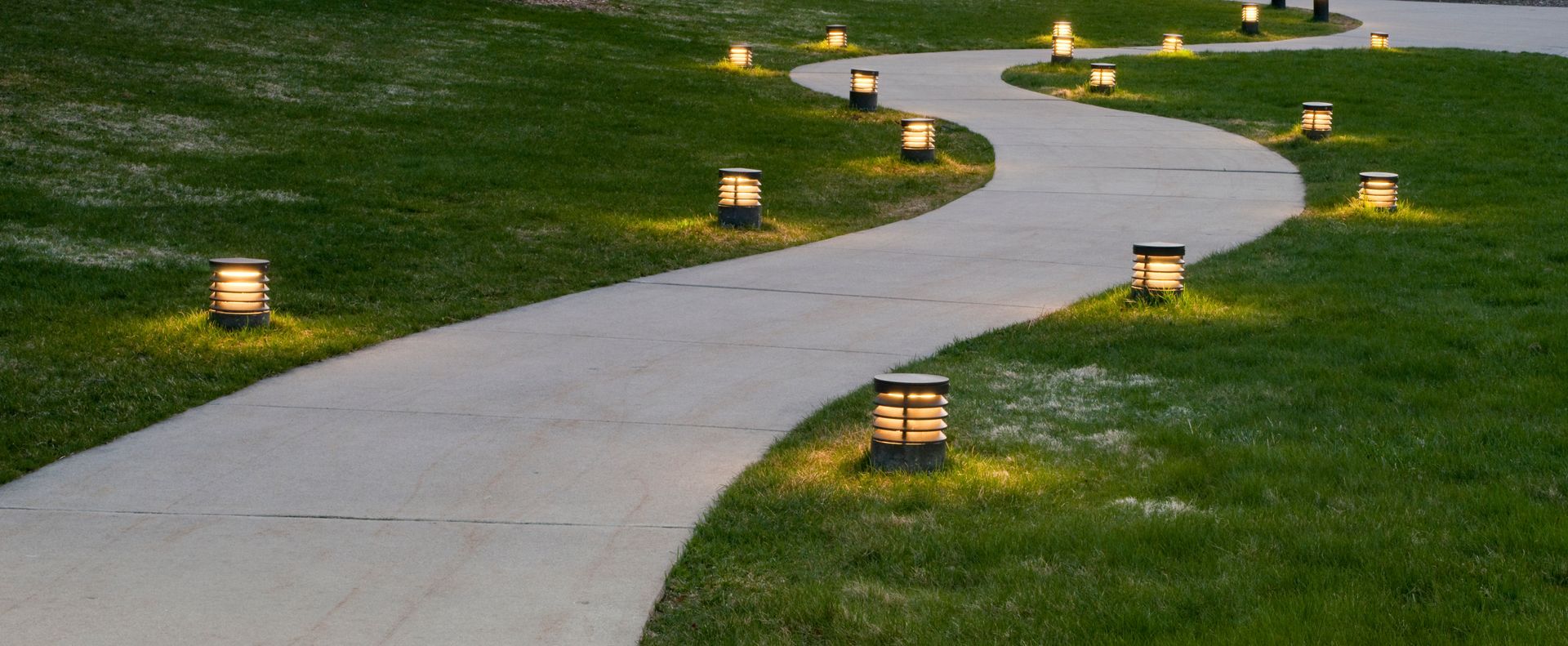 A path with lights on the side of it going through a grassy field.