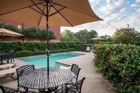 Pool area with round table, chairs, beige umbrella, hedges, and chaise lounges.