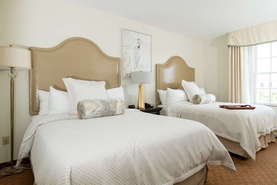 Two beds in a hotel room with white bedding, beige headboards, and a window.