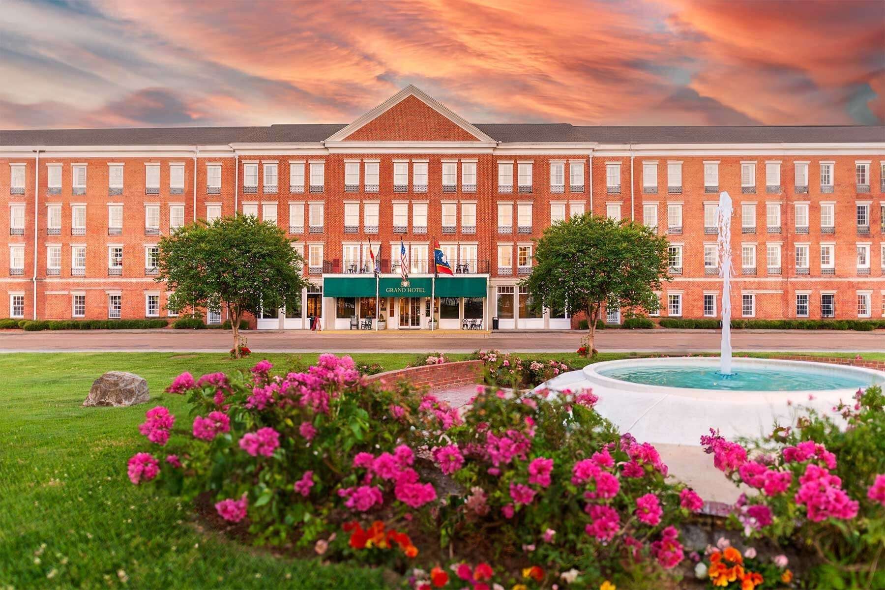 Hotel exterior with flowers and fountain. Brick building, green lawn, and colorful sunset.