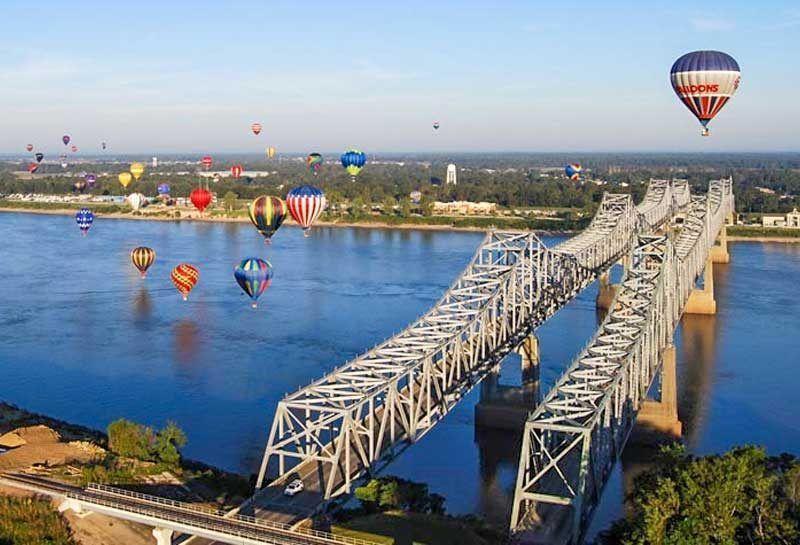 Hot air balloons over a river bridge, with trees and buildings in the background.