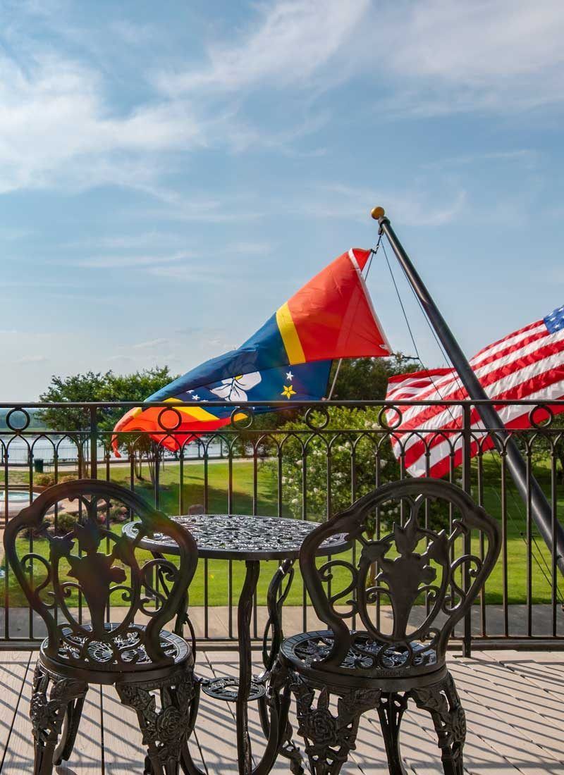 Two ornate chairs and table on a patio overlooking water, flags waving in the breeze.