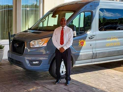 Man in tie stands beside a gray passenger van. Exterior setting, well-dressed.