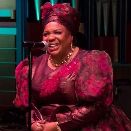 Woman singing at a microphone, wearing a red headwrap and dress. Inside, with musical instruments.