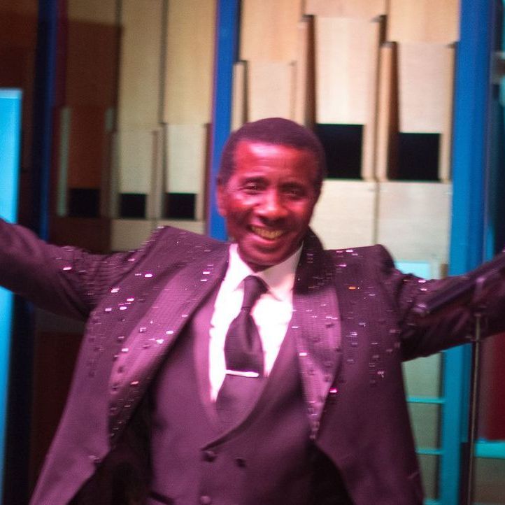 Smiling Black man in a decorated black suit with arms outstretched, performing on stage with a warm, wooden backdrop.