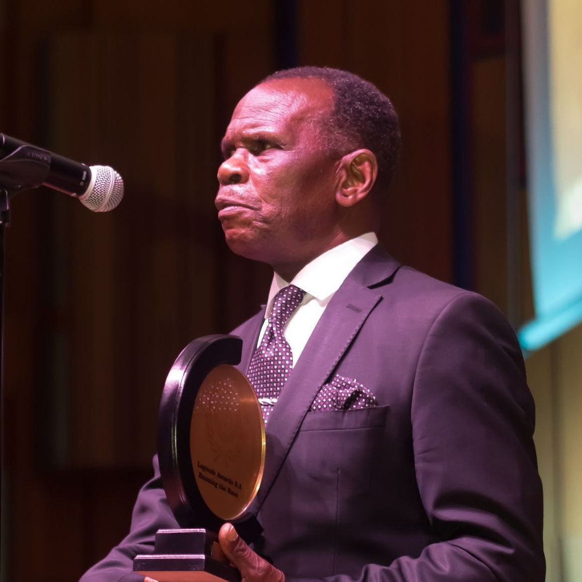 Man in a suit holding an award, speaking into a microphone on a stage.