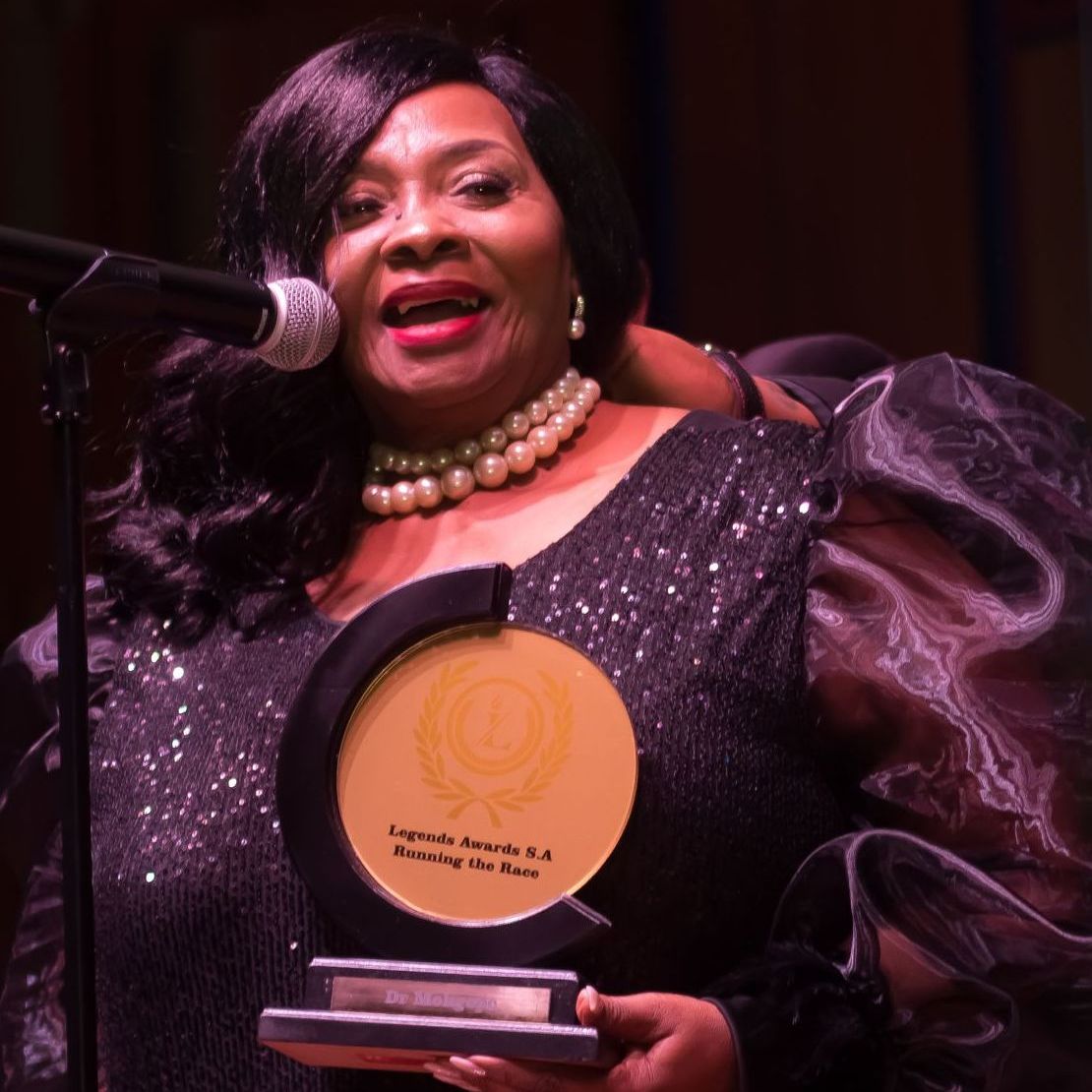 Woman in formal wear holding an award, smiling at a podium with a microphone.