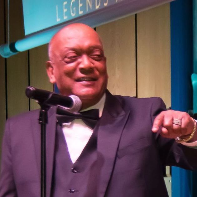 Man in suit speaking at microphone, smiling, gesturing with his right hand.