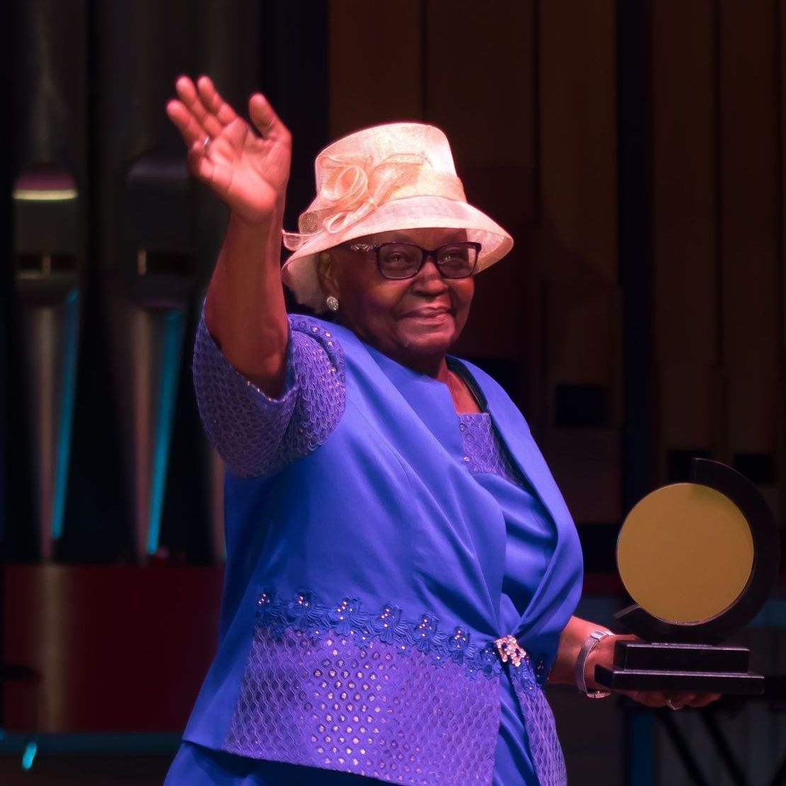 An older Black woman in blue waves, holding an award, and wearing a hat.