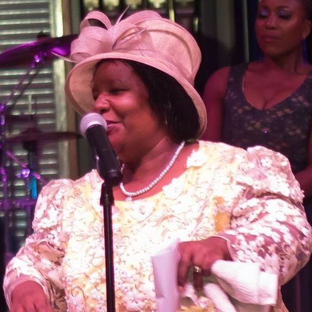 Woman in a yellow dress and hat singing into a microphone on stage.