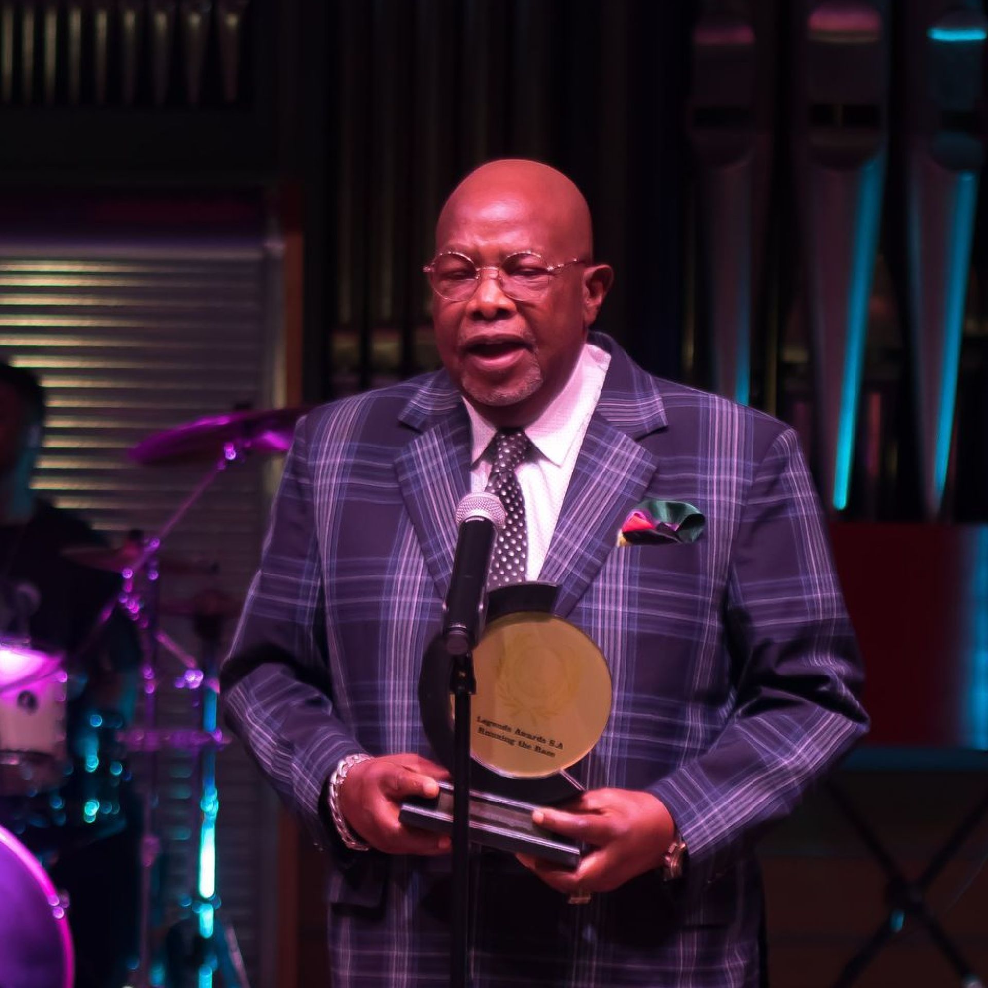 Dr Elijah Maswanganyi holding an award, speaking into a microphone on a stage.