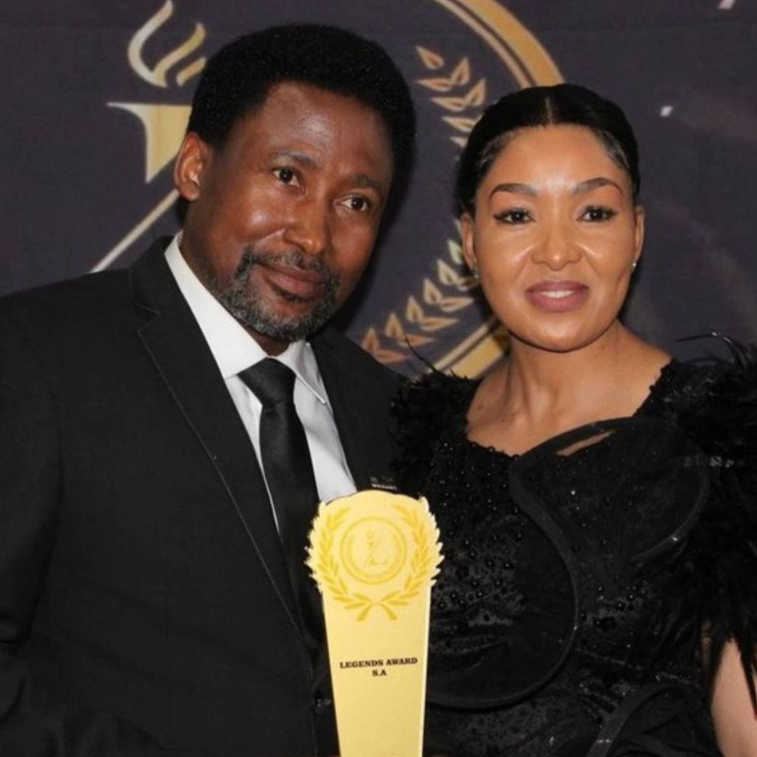 Man and woman at awards ceremony; man holds gold award, both smile.