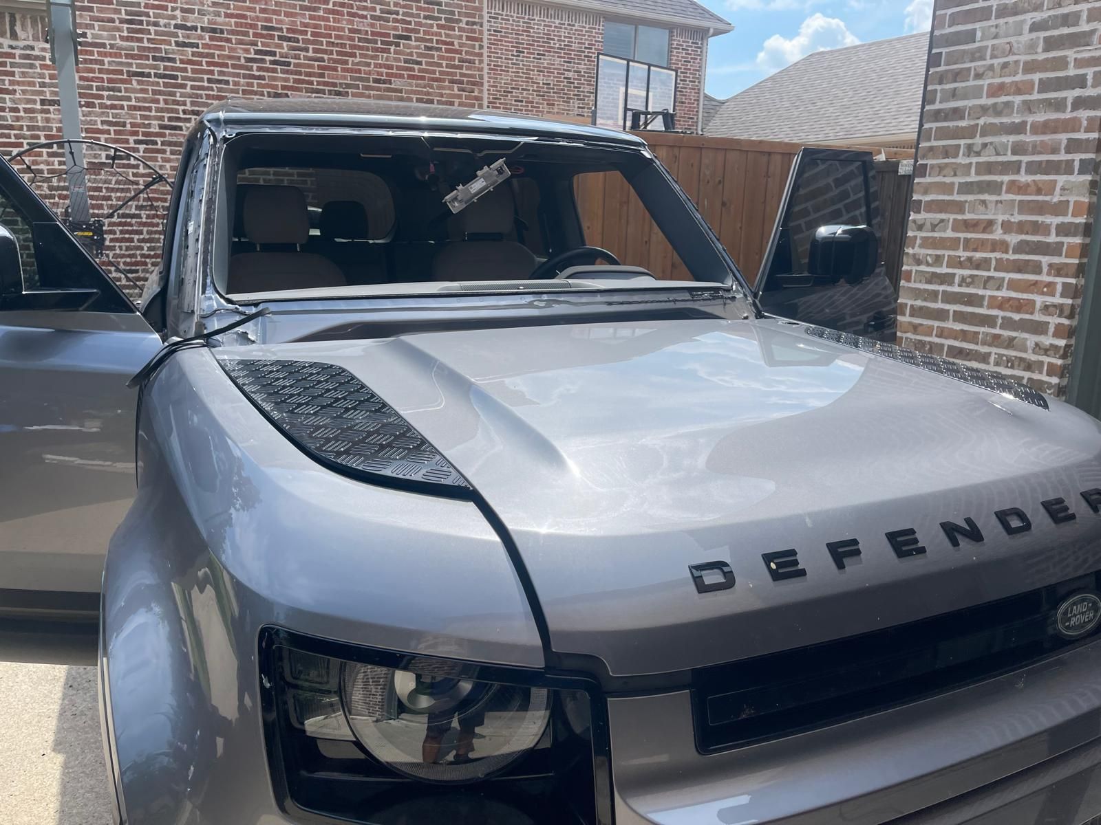 Gray Land Rover Defender with a broken windshield; parked outside a brick house.