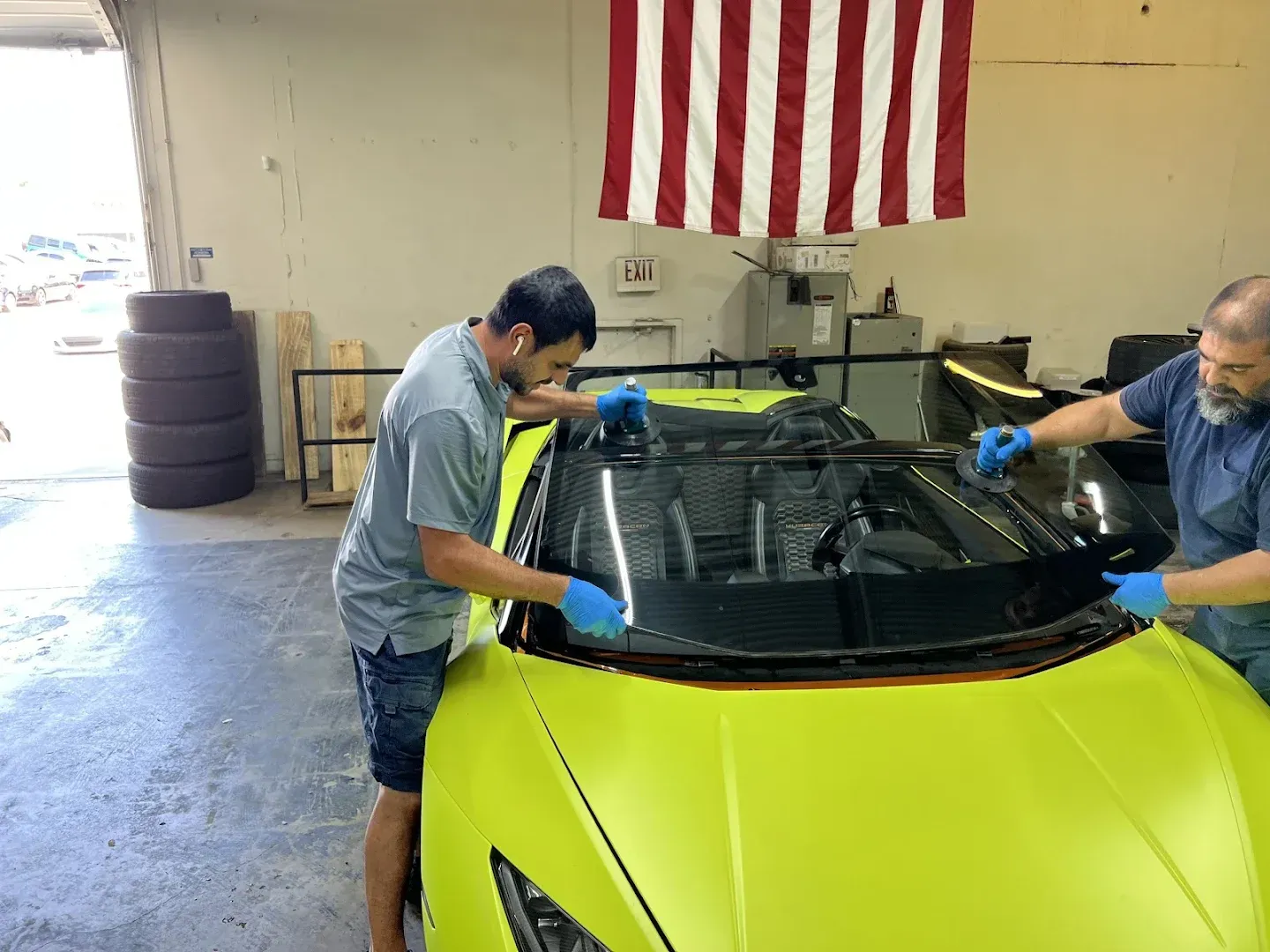 Two men installing a black windshield on a bright yellow sports car indoors, under an American flag.
