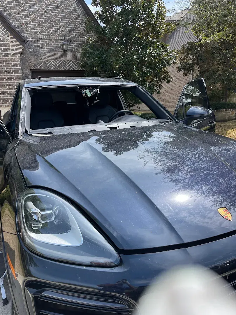 Dark gray Porsche SUV with shattered windshield, damaged. Outdoors, in front of a building.