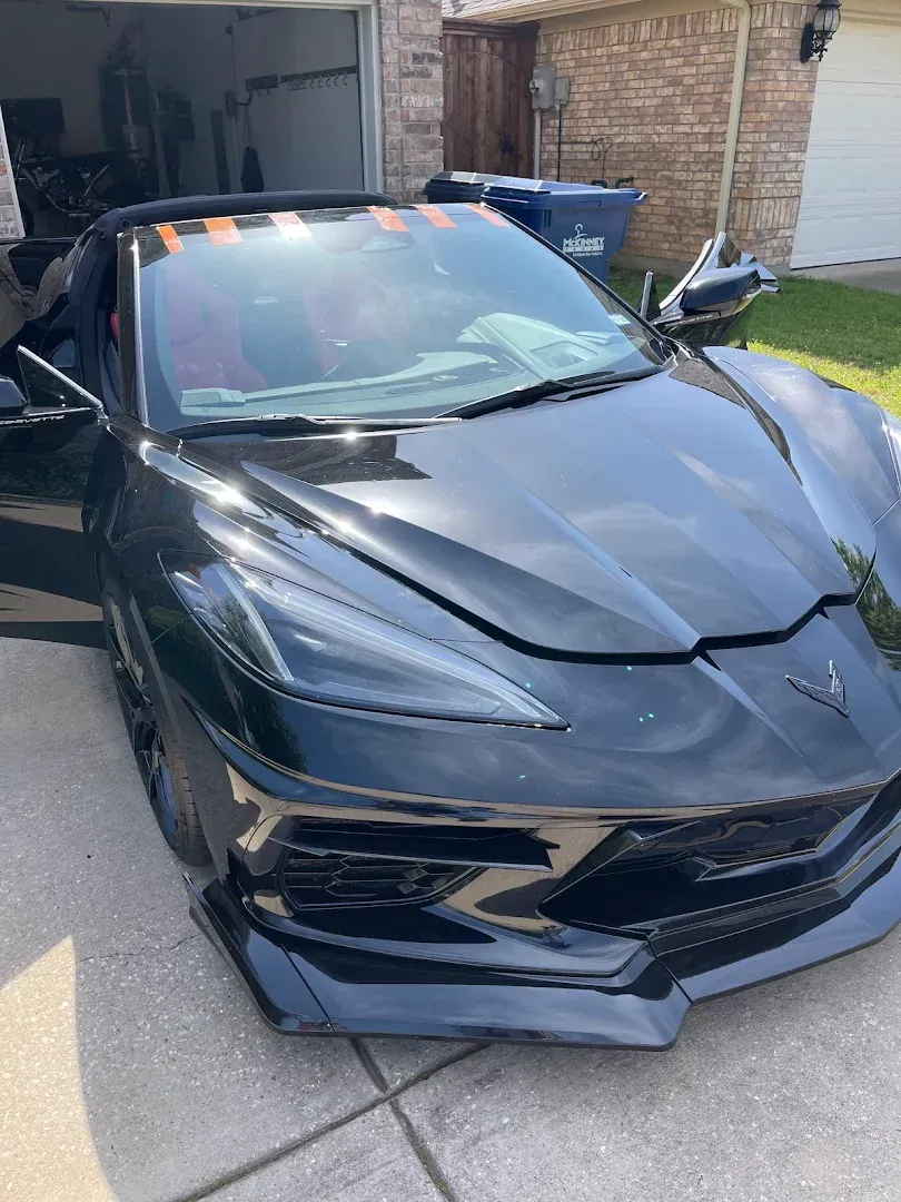 Black Corvette convertible parked on a concrete driveway. The driver's side door is open.