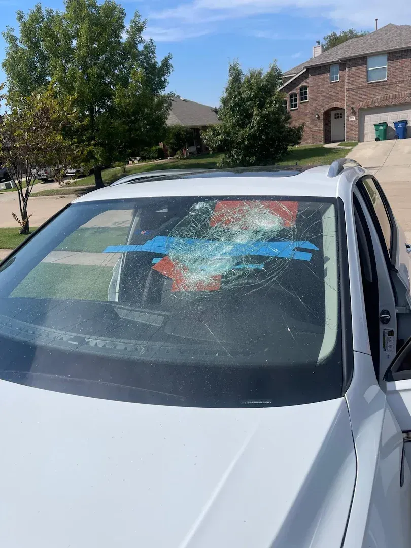 White car with shattered windshield, parked on a suburban street under a blue sky.