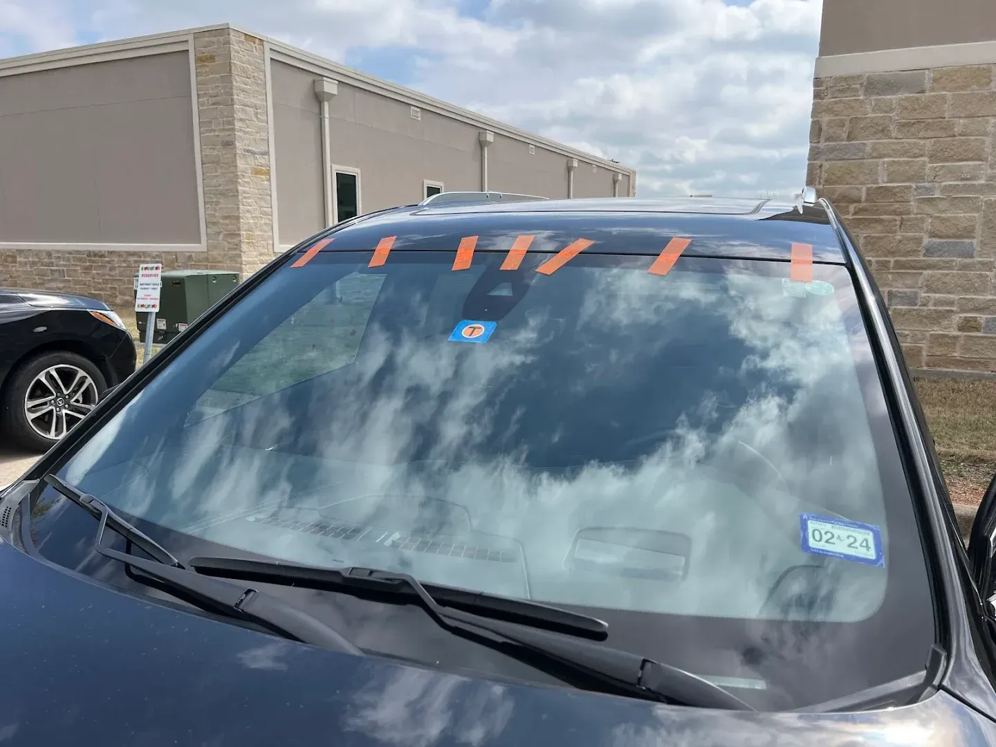 Black car with cracked windshield, taped, reflecting cloudy sky.