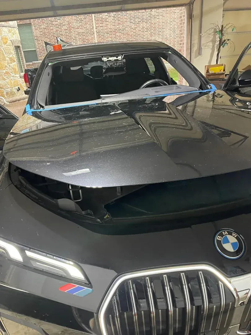 Black BMW car with windshield being replaced in a garage. Hood open, blue tape visible.