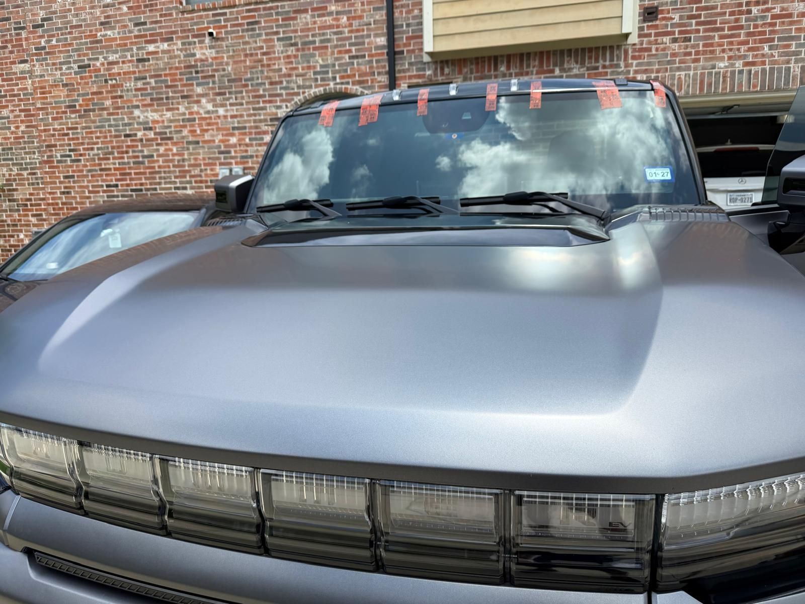 Silver car front parked in front of a brick building, windshield reflecting clouds.