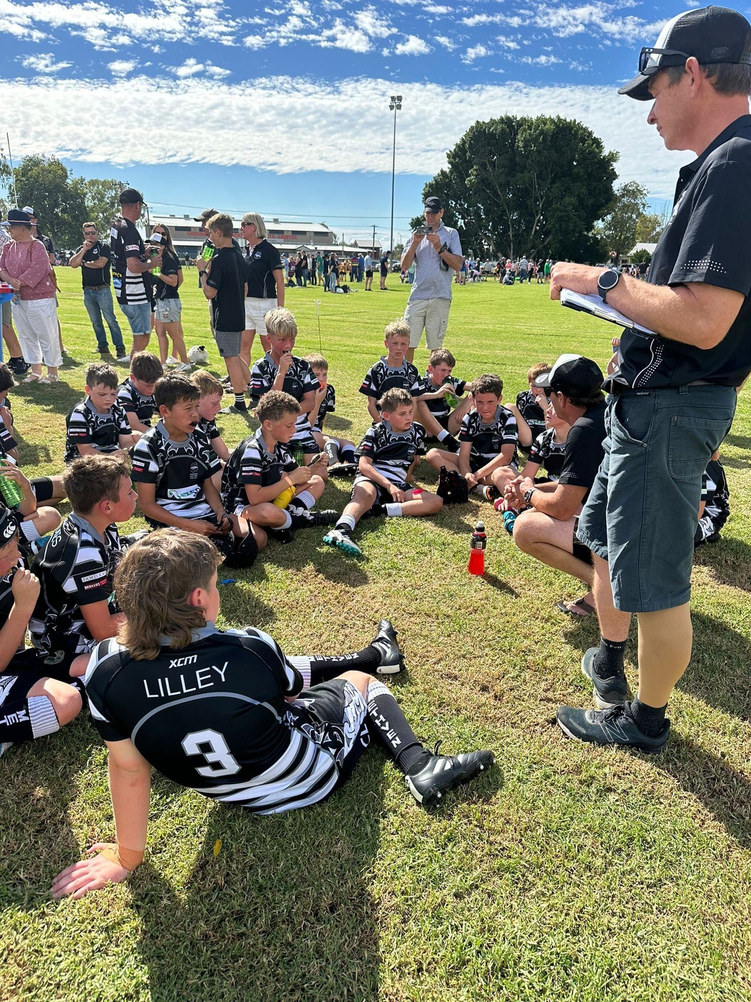 A group of young boys are sitting on the grass while a man talks to them