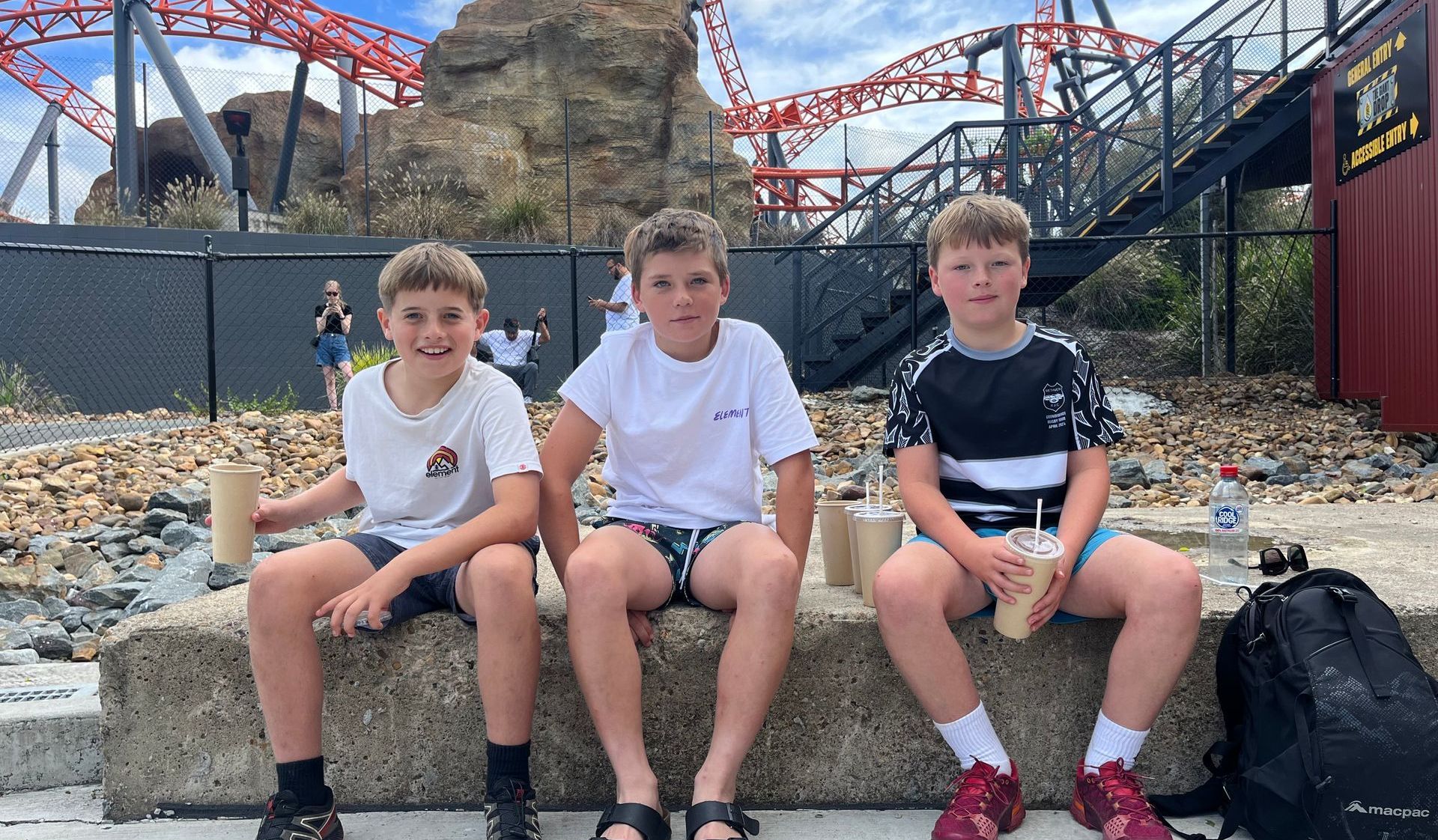 Three young boys are sitting on a stone wall in front of a roller coaster.