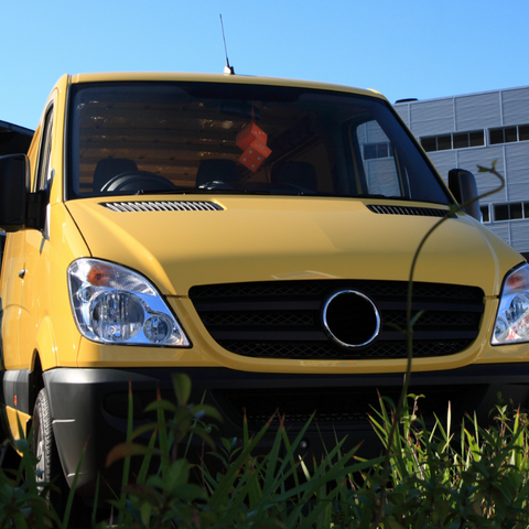 Yellow cargo van parked in front of a building on a sunny day. Tall grass is in the foreground.