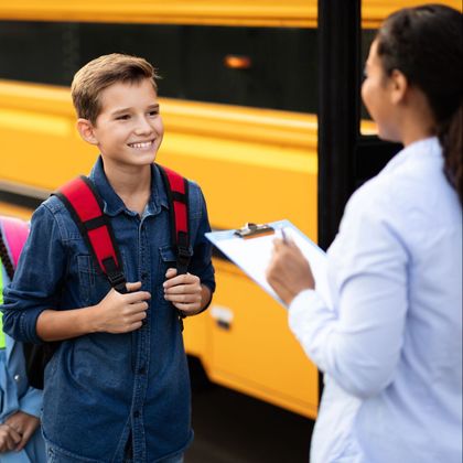 Boy with backpack smiles at a woman with a clipboard near a yellow school bus. They appear to be in a school setting.