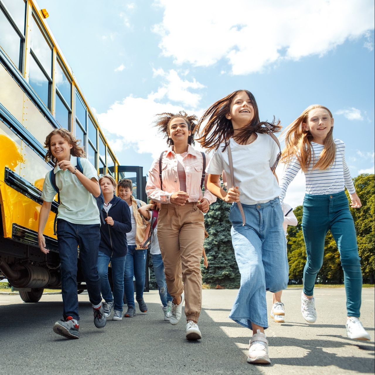 Children excitedly run away from a yellow school bus on a sunny day. They wear casual clothes and backpacks, smiling.