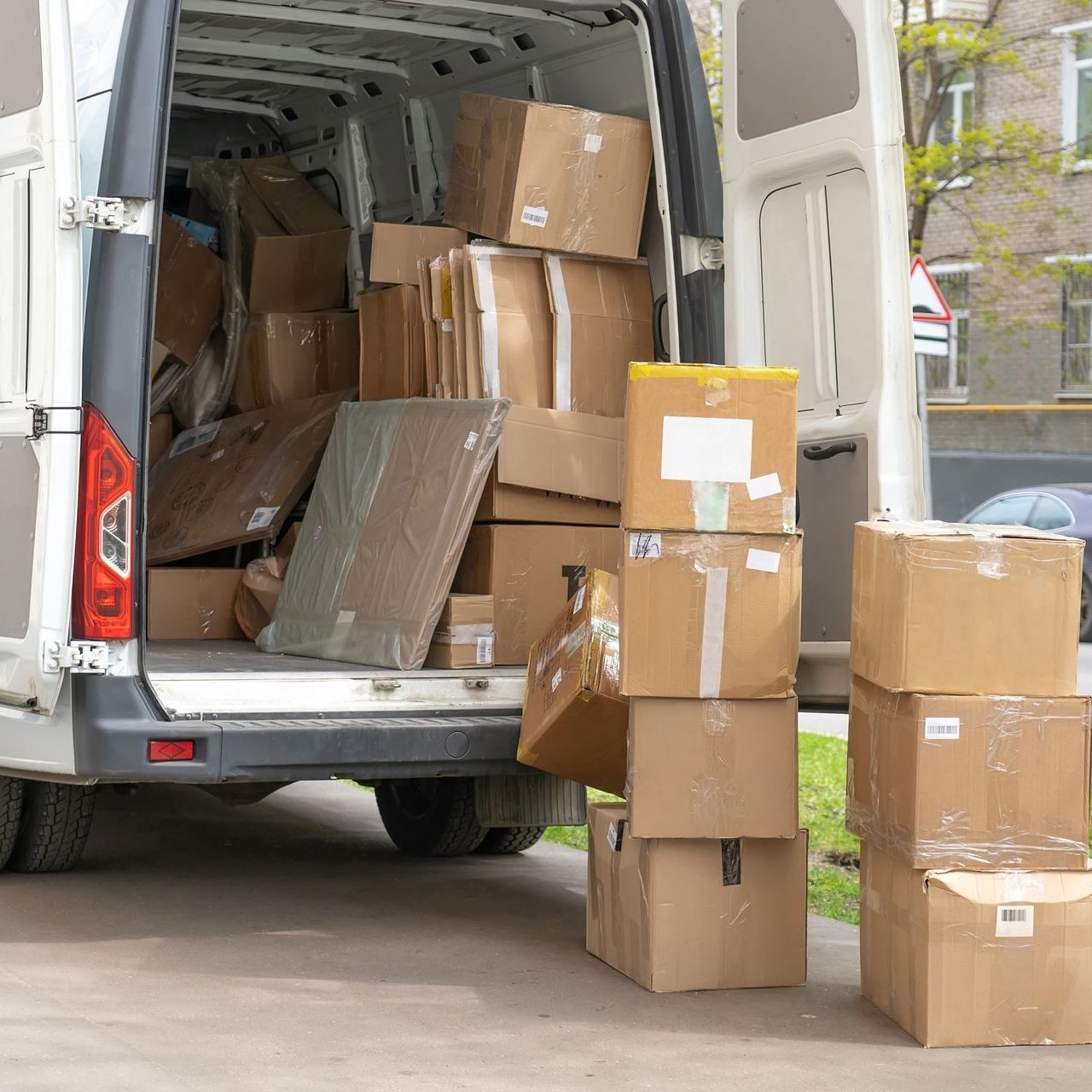 A white van with its back doors open, packed with cardboard boxes and more boxes stacked outside on the sidewalk.