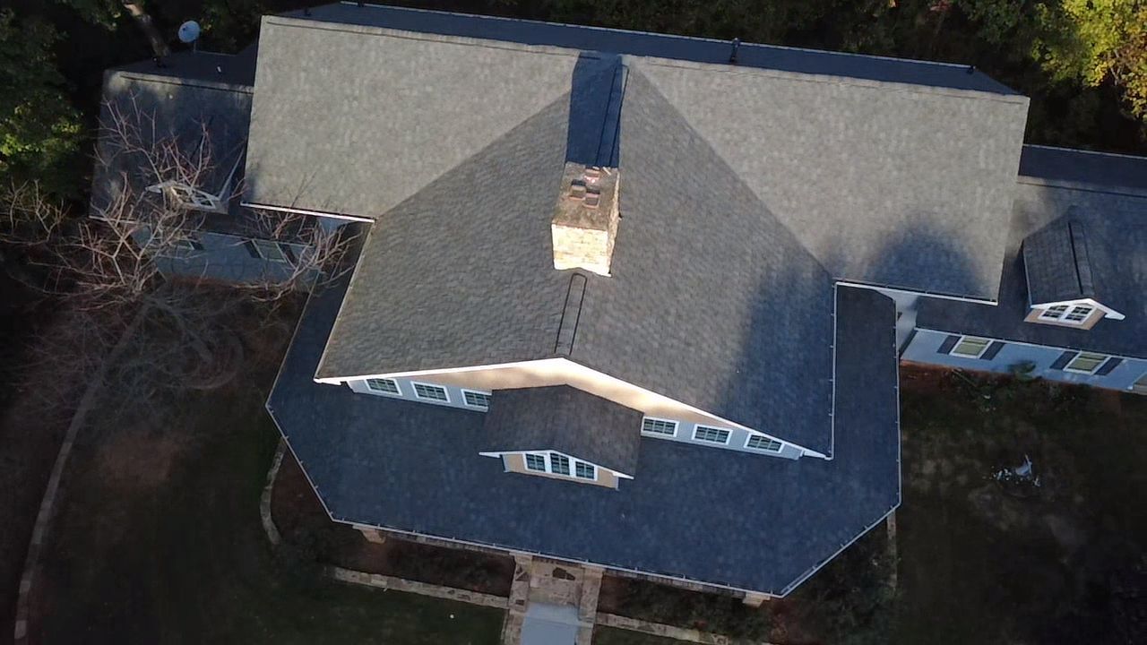 An aerial, top-down view of a house with a grey shingled roof, featuring a central chimney and surrounding greenery.