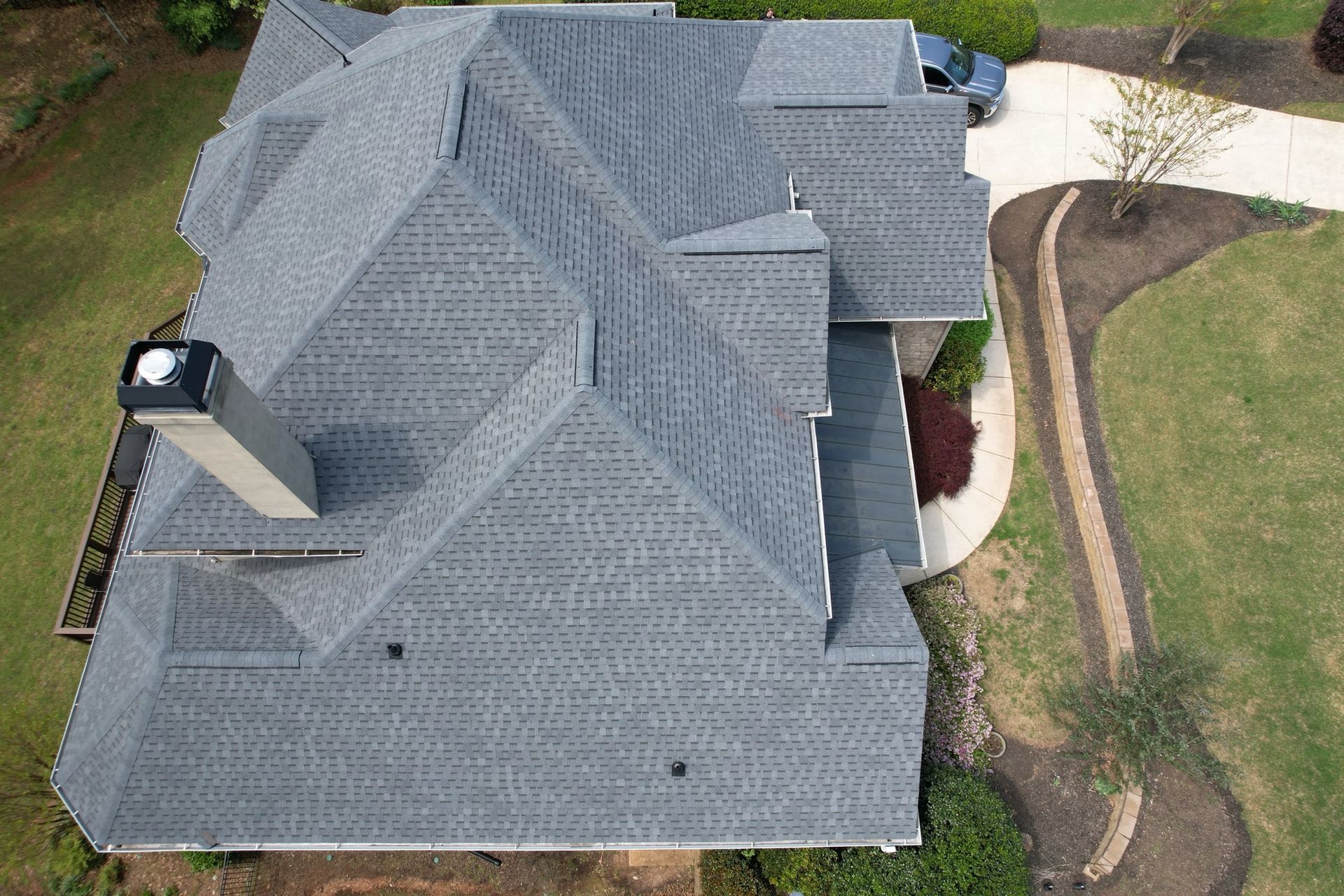 An aerial view of a gray shingled residential roof with a chimney, surrounded by grass and a curved walkway.