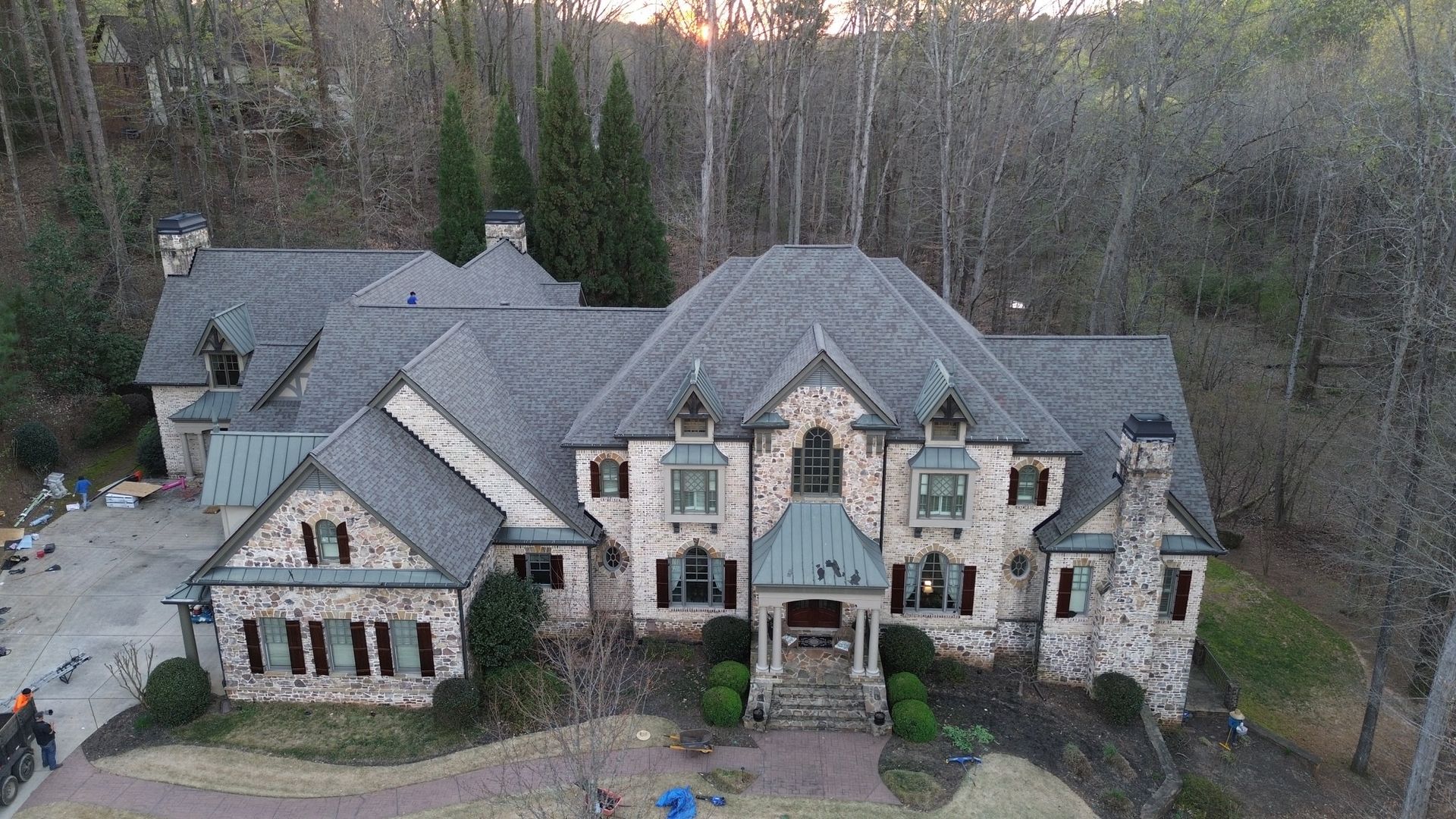 An aerial view of a large, multi-story stone estate house with a grey shingled roof, surrounded by trees.