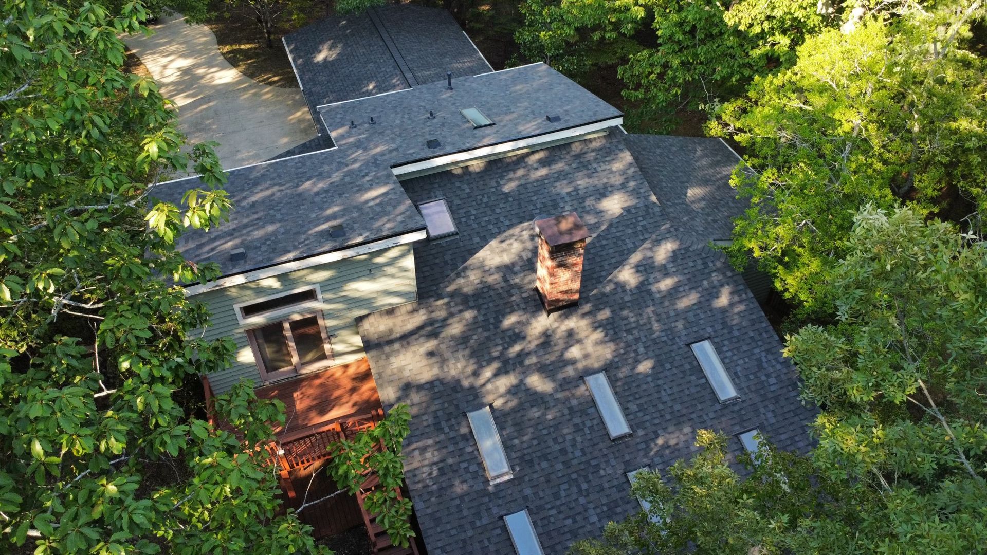 An aerial view of a dark shingled house roof with a brick chimney and several skylights surrounded by dense green trees.